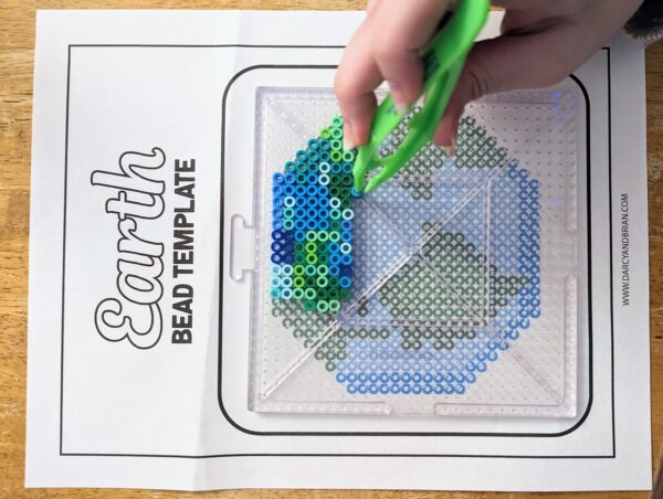 Earth bead pattern printed out and child adding beads with a green tweezer onto pegboard.