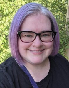 Headshot of a white woman with short purple and gray hair wearing purple glasses and standing in front of a tree.